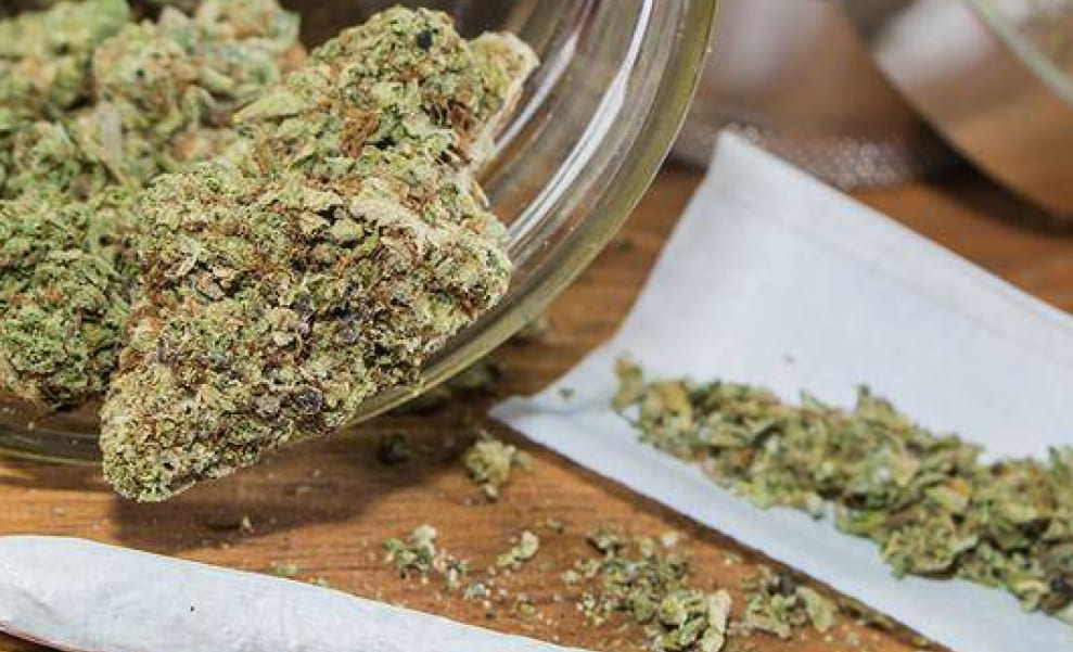 A clear glass bowl filled with cannabis buds spilling onto a wooden surface next to rolling papers and loose ground cannabis.