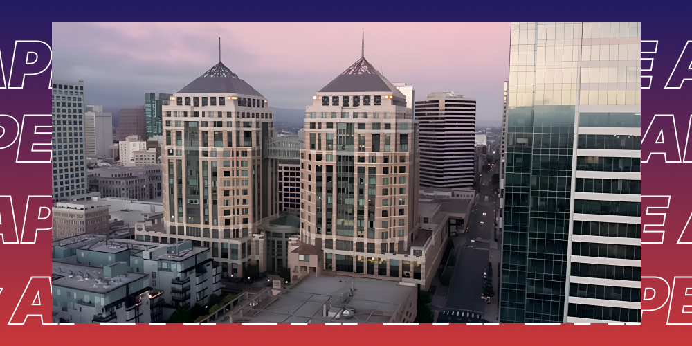 An aerial view of two distinct, ornamental twin towers in downtown Oakland, CA, set against a backdrop of other skyscrapers during a pink and blue twilight. The image is framed by a colorful, repeating 'APE' pattern.