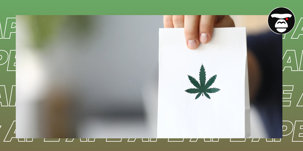 A hand holding a plain white paper bag with a dark green cannabis leaf logo printed on it.