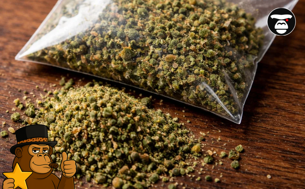 Ground green cannabis flower known as shake or pre-ground bud sitting next to a plastic baggie on a wooden table.